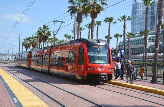 Red San Deigo light rail train at a stop on a sunny day and palm trees.