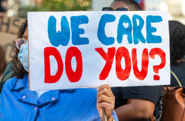 Image of a masked woman holding a sign with text: We care, do you?