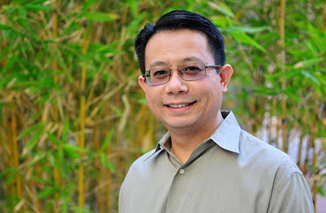 Smiling man with dark hair, glasses, gray button down shirt, standing in front of leafy bamboo.