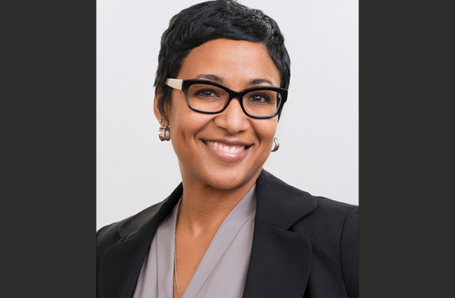 Dr. Amani Allen, a smiling Black woman with short black hair, wearing black glasses a grey blouse, and a dark blazer