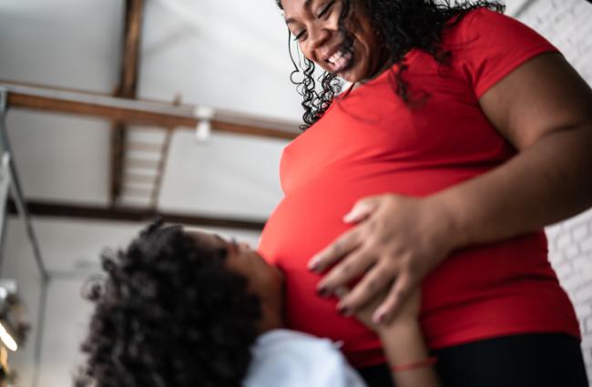 Young Black-presenting person putting their hands on the pregnancy bump of a Black- and female-presenting person.