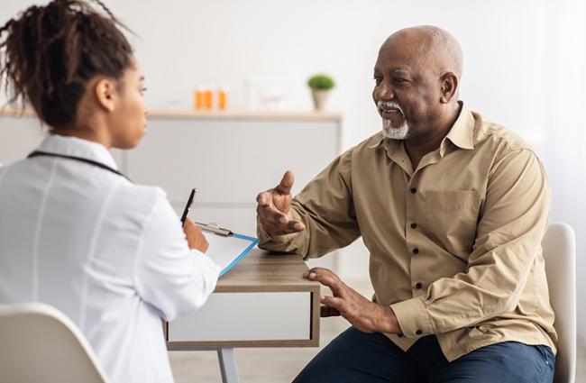 Black senior man sitting down and talking to a Black female doctor who is filling out a form while listening to an elderly patient