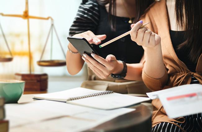 Two female-appearing people with manicured nails are looking at a phone over a desk. Underneath there are white notepads, and in the background is an old fashioned balance/weigh scale, insinuating some sort of law office.