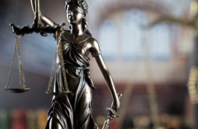 A brass-colored metal female statue holding up a balancing scale with her right arm, and in her left arm she is holding a long sword pointed toward the ground. The background is a blurred out library with a book shelf.