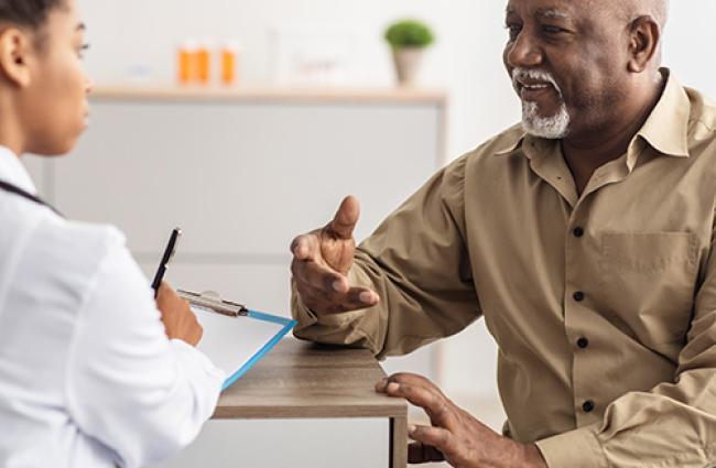 Black senior man sitting down and talking to a Black female doctor who is filling out a form while listening to an elderly patient