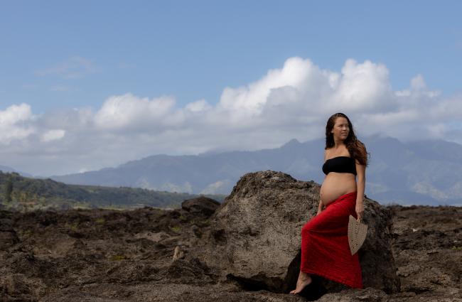 Pregnant Native Hawaiian person wearing a long red skirt and black bandaue standing on rock with mountains in the background