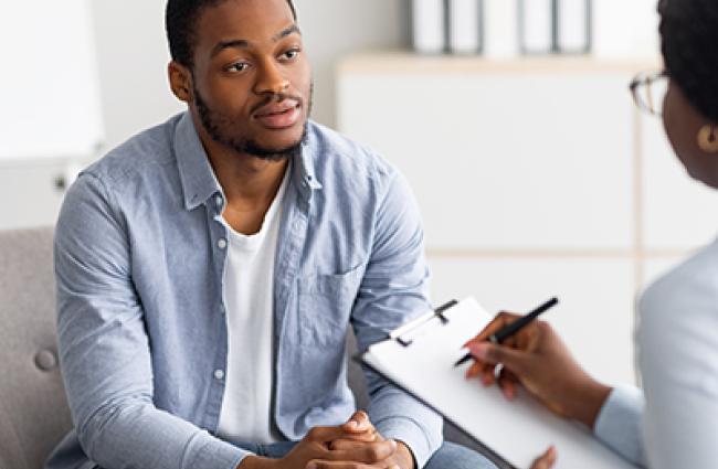 A serious-looking Black young man sits and talks to a doctor who holds up a clipboard