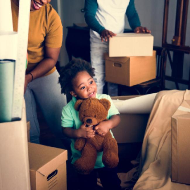 A smiling little girl holding a teddy bear while parents move boxes