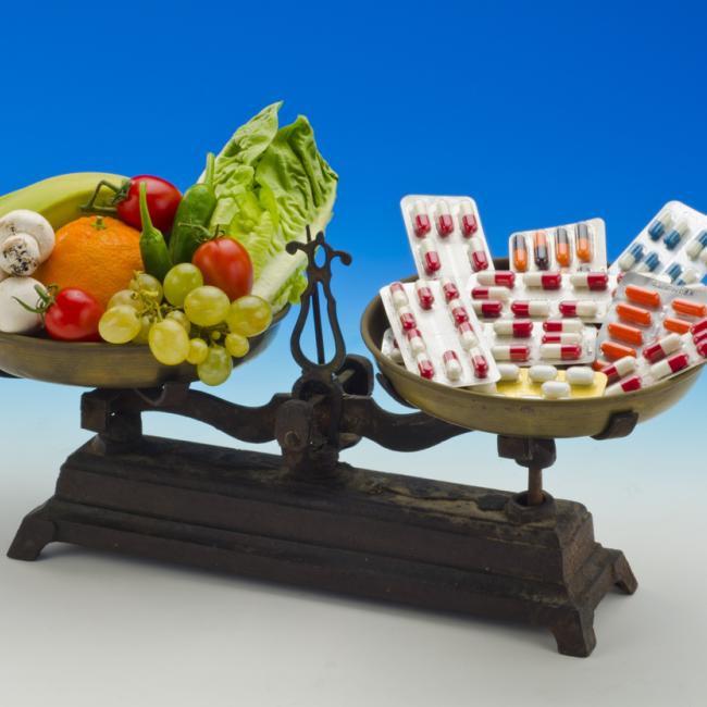 Image of a two-armed scale with vegetables and fruits on the left hand side and colorful pills on the right hand side.