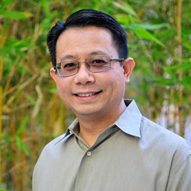Smiling man with dark hair, glasses, gray button down shirt, standing in front of leafy bamboo.