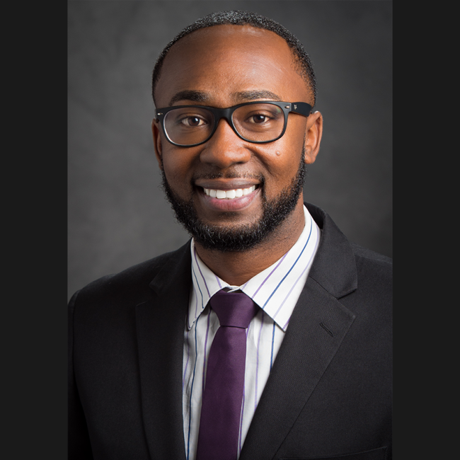 Charles H. Lea III, a smiling Black man with short cropped hair, glasses, and a short beard and mustache. He's wearing a striped dress shirt, dark plum colored tie, and a dark suit jacket.