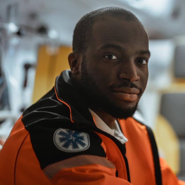 In the foreground, there’s a young Black man wearing an emergency medical response orange jacket and looking into the camera, and a blurred background of the inside of an ambulance.