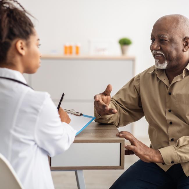 Black senior man sitting down and talking to a Black female doctor who is filling form listening to elderly patient