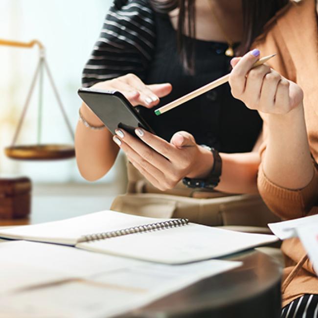 Two female-appearing people with manicured nails are looking at a phone over a desk. Underneath there are white notepads, and in the background is an old fashioned balance/weigh scale, insinuating some sort of law office.