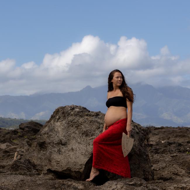 Pregnant Native Hawaiian person wearing a long red skirt and black bandaue standing on rock with mountains in the background