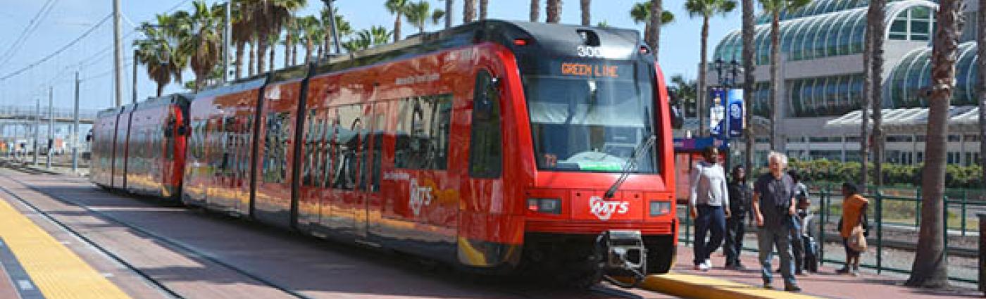 Red San Deigo light rail train at a stop on a sunny day and palm trees.