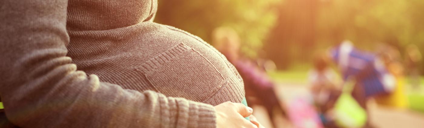 Pregnant person wearing a brownish-grey top with their arms wrapped around their stomach