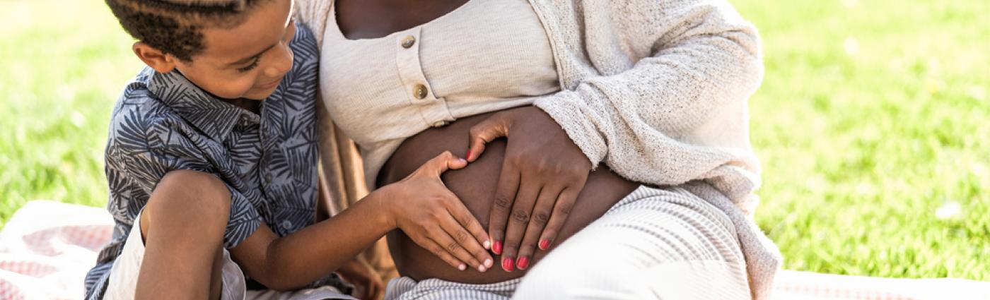 Black pregnant person sitting in a grassy area with a young Black child, both of whom have their hands on the pregnant person's abdomen