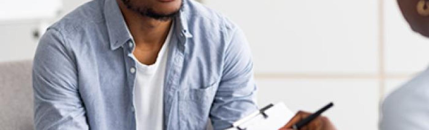 A serious-looking Black young man sits and talks to a doctor who holds up a clipboard