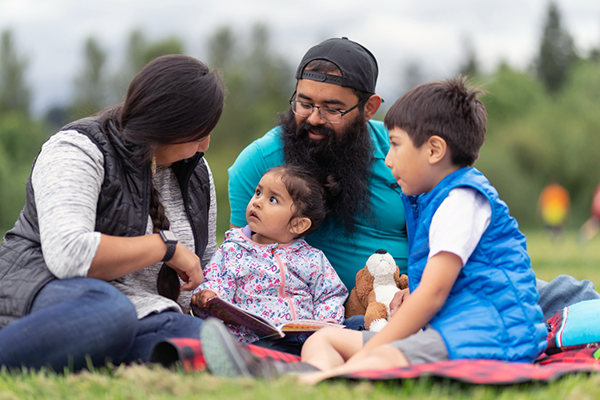 An Indigenous mom and dad hanging out at the park and reading a books with their two young children on a warm summer afternoon. They're all sitting on the grass together.