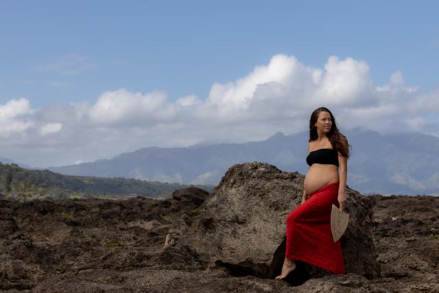 Pregnant Native Hawaiian person wearing a long red skirt and black bandaue standing on rock with mountains in the background