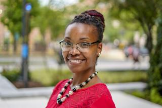 Dr. Jeana Morrison, a smiling Black woman with braided hair twisted into a bun at the back of her head. She's wearing glasses, a beaded necklace, and a red patterned top. She's standing in front of a blurred background of what looks like a park or part of a college campus.