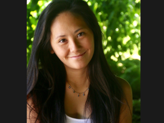 Elissa Carey, a smiling Chinese American woman with long dark hair. She's wearing a dangly beaded necklace and white top. She's sitting in front of a green leafy background.
